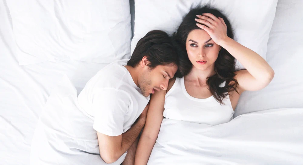 Annoyed woman lying awake in bed next to her sleeping partner